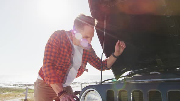 Stressed caucasian man looking at broken down car with open bonnet on sunny day at the beach