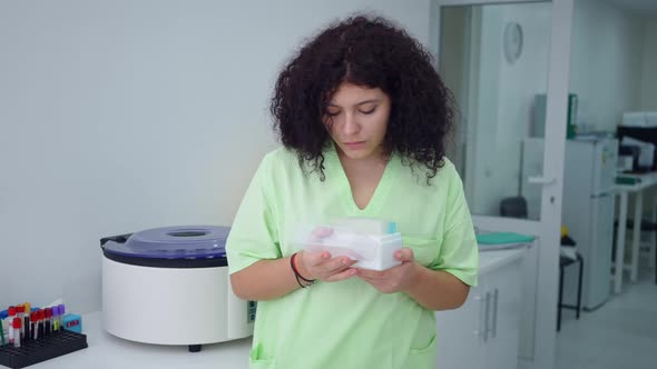 Concentrated Plussize Lab Assistant Examining Test Tubes Box and Looking at Camera Smiling