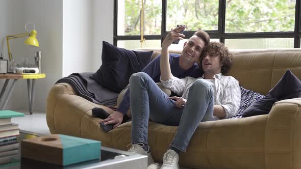 Happy Young Gay Couple Sit on Couch in Living Room Doing Video Call with Family Friends Using Cell