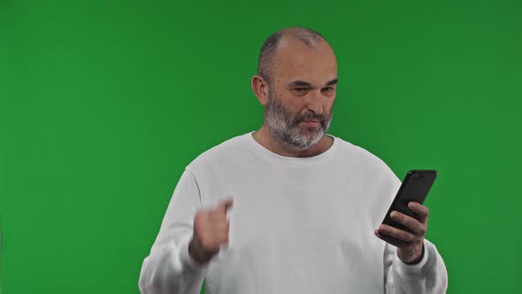 Happy Senior Man Showing Yes Gesture Holding Smartphone on Isolated Green Background