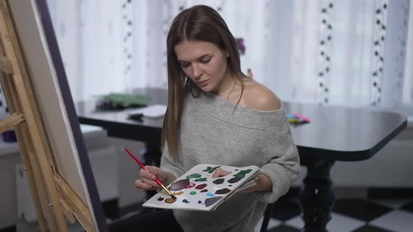 Medium Shot Portrait of Young Slim Woman Mixing Colors on Painting Palette Shaking Head Sighing