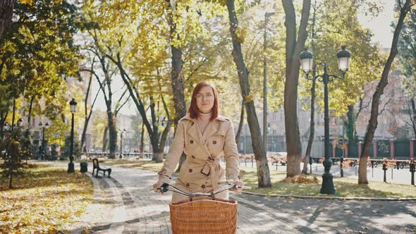 Young Redhead Female is Riding Cruiser Bike By Tiled Path of Autumn City Park