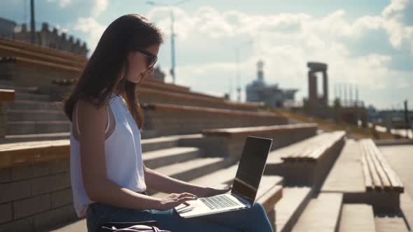 Smiling Woman Student Sitting on Bench Outdoors at the Street, Using Laptop Computer, Use Mobile