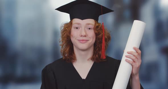 Young Student in a Robe and Hat Holds a Diploma Paper Scroll in His Hands, a Red-haired Teenager