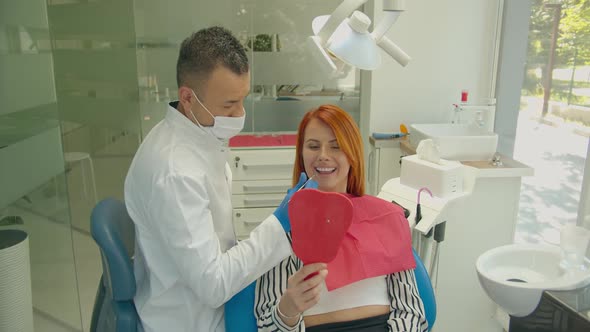 Professional Male Dentist Show Beautiful Teeth in Mirror To Happy Woman Visitor. Positive Red-haired
