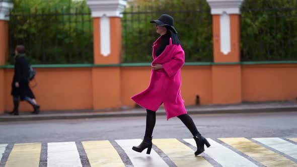 A Brunette Woman in a Pink Coat and a Black Hat Runs Along an Asphalt Road Against the Background of