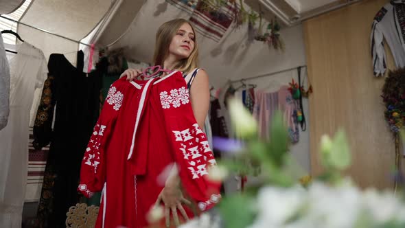 Smiling Teenage Ukrainian Girl Choosing Traditional Embroidered Dress in Handcraft Shop Indoors