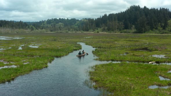 Drone shot of two people paddling a canoe through a small waterway surrounded by grass and forest