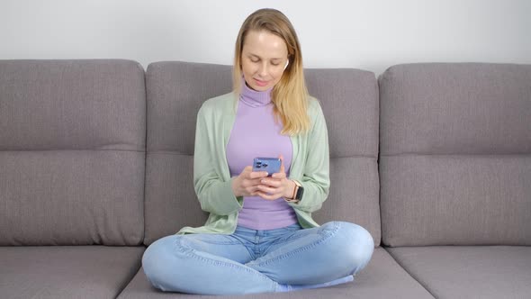 Blonde woman using mobile phone and headphones on sofa in living room in 4k video