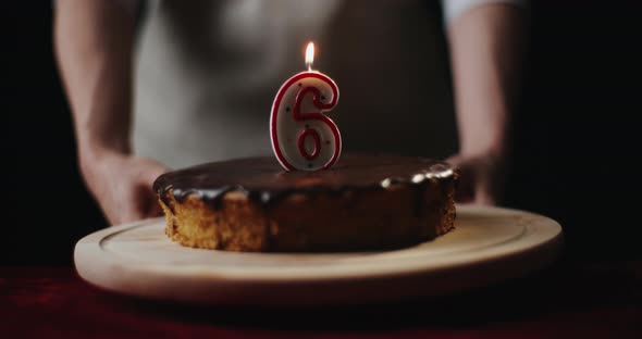 Young Woman Wearing Kitchen Apron Puts Homemade Chocolate Cake or Pie with Number 6 Burning Candle