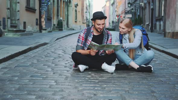 Trendy Couple of Tourists Using Map, Sitting on Pavement and Admiring Historical Surroundings