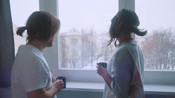 Happy Young Multiracial Couple Drinking Morning Coffee in Front of the Window and Watching Park