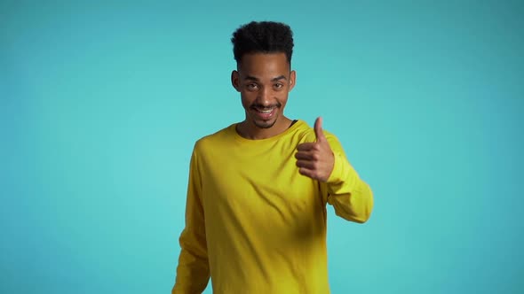 Handsome black man in yellow wear on blue studio background smiles to camera and gives thumbs up