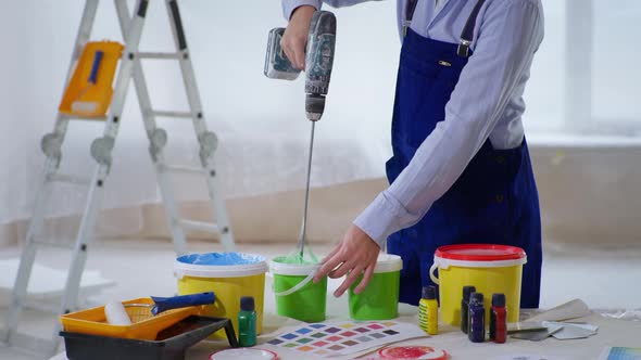 Male Construction Worker in Work Clothes and Helmet Mixes Paint in Bucket Using Drill To Paint Walls