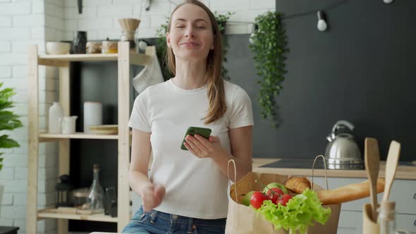 Woman Stands in the Kitchen Near a Paper Bag Full of Fresh Food and Uses a Smartphone App to Deliver