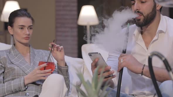 Couple At Shisha Bar. Man Smoking Hookah And Using Phone