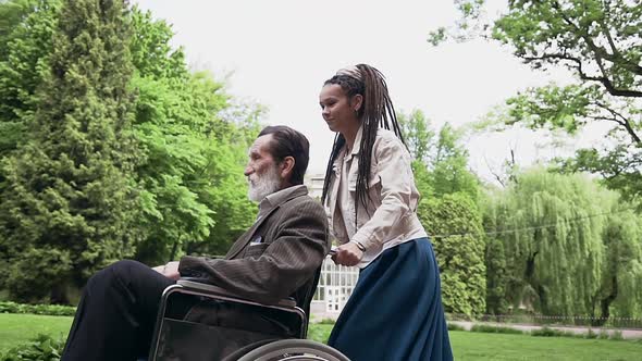 Young Girl with Dreadlocks Helping Senior Positive Sedentary Grandfather in Wheelchair in the Park