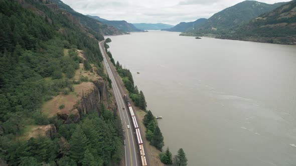 Aerial rise above train and highway traffic traveling next to Columbia River.