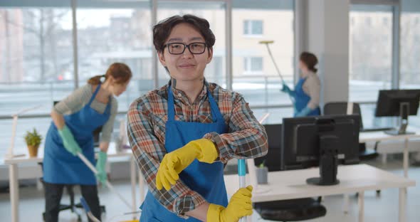 Asian Janitor in Glasses and Uniform Smiling at Camera with Colleagues Working on Background