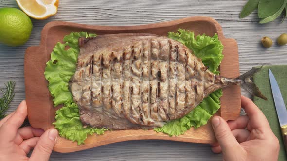 Hands are Placing a Wooden Plate with a Delicious Juicy Grilled Mackerel Fish