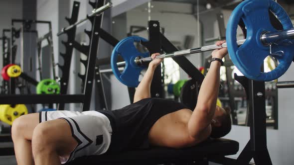 Athletic bodybuilder man trains in gym with barbell lifting it, side view.