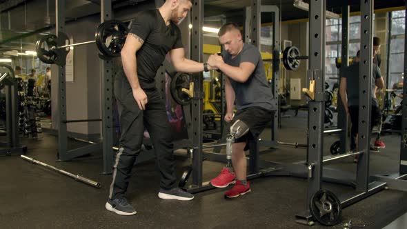 Instructor Giving a Hand To Young Man Exercising with Prosthetic Leg in Gym