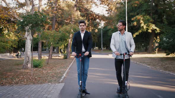 Two Handsome Young Businessman Riding an Electric Scooters on the Road in City Park