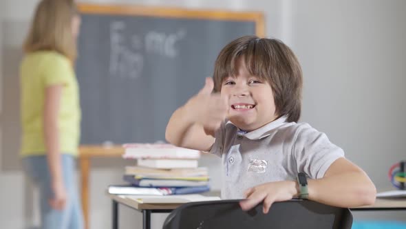 Cute Excited Schoolboy Turning To Camera Showing Thumb Up with Blurred Classmate Writing on School