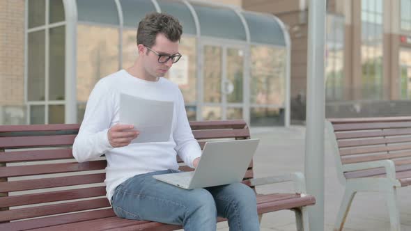 Young Man Celebrating Success While Reading Documents and Working on Laptop Outdoor