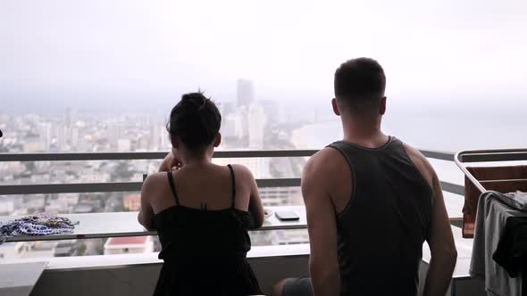 Back View of Young Couple Sitting on Balcony in High Building with View of City