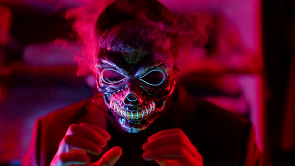 a Man in a Scary Glowing Death Mask Lets Out Puffs of Smoke and Joins Hands in a Neon Flashing Light