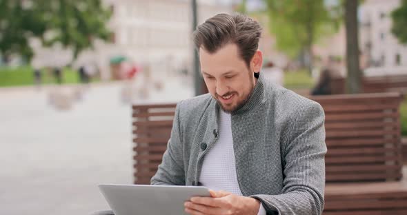 Young Man with Mustaches and a Beard Sitting on a Bench in the Square Typing on the Laptop Keyboard