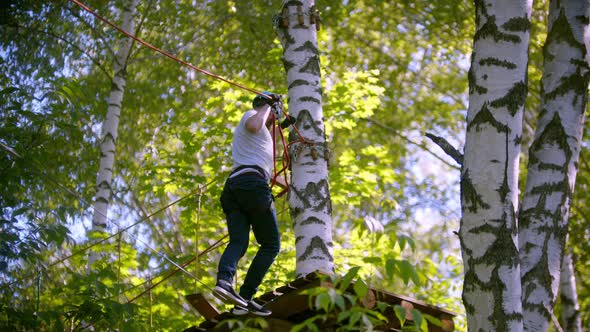 A Man in Helmet Carefully Walks on the Rope Suspended in the Air Between Trees in the Forest