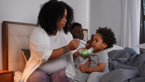 Young African American Mother Feeding Cute Son with Spoon As Blurred Father Reading Lying in Bed at