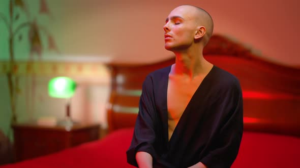 Sad Extravagant Male Queer Sitting on Bed in Red Light and Turning to Camera