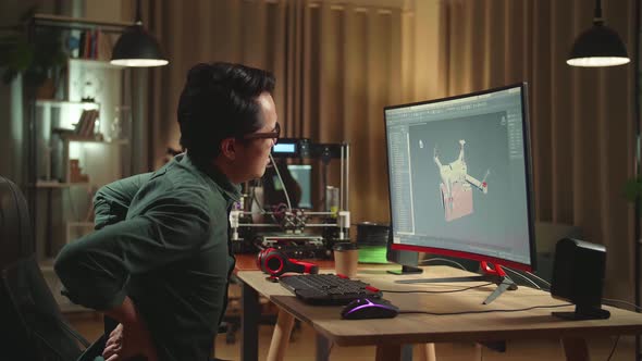 Asian Engineer Paining His Back While Work On Computer And 3D Printer, Screen Shows 3D Drone Model