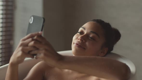 Young Cute African American Lady Relaxing in Bath at Home Web Surfing Social Media on Smartphone