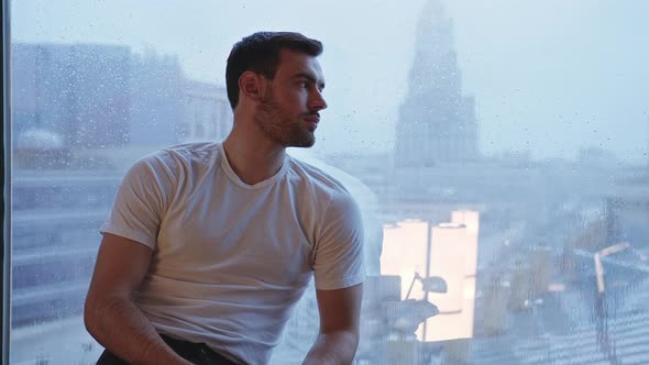 A Joyful Young Man Sits By the Window on a Rainy Day