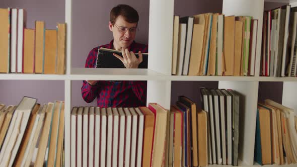 A Man Wearing Glasses is Reading a Book in the Library Behind the Bookshelves