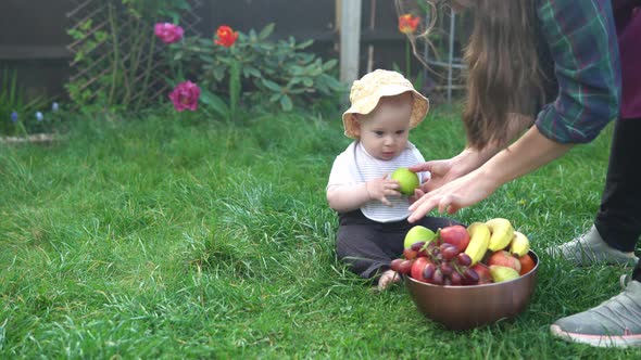 Small Newborn Child in Summer Panama Hat Sit on Grass Barefoot in Bib with Big Bowl of Fresh Fruit