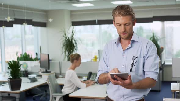Young Handsome Businessman Holding Tablet Showing Thumb Up Coworker Working on Background