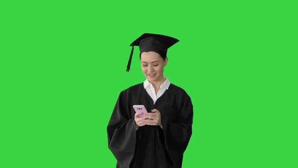 Smiling Female Graduate in Mortarboard Sharing Happy News on Her Phone While Walking on a Green