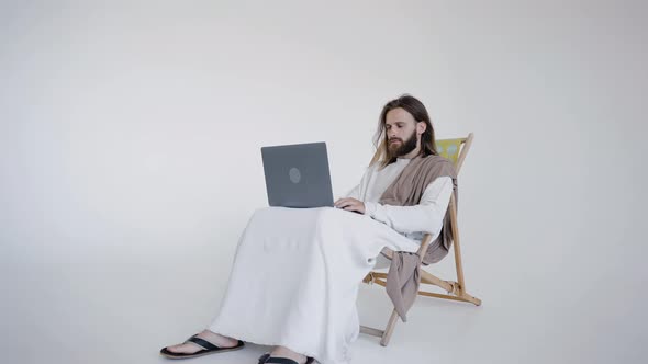 Jesus Sits in a Schizlong and Works at a Computer on a White Background