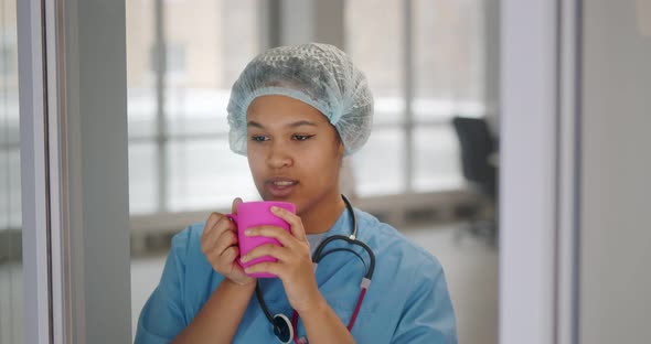 Young African Nurse in Uniform Relaxing Near Window with Cup of Coffee and Smiling