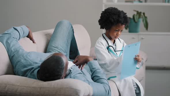 Serious Child Pretends to Be Doctor in Medical Costume Sitting Writing Symptoms on Tablet Father