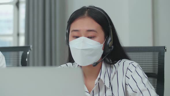 Close Up Of An Asian Woman Call Centre Agent Wearing Headset, Mask And Speaking To A Customer