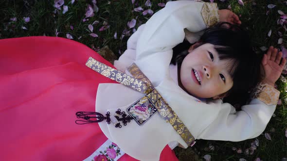 Korean Girl Child in a National Costume Lies on the Back in a Garden with Cherry Blossoms in Spring