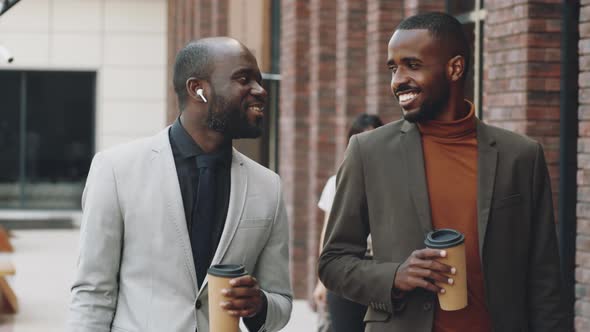 Two Black Businessmen Chatting and Walking on Street with Coffee