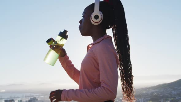 African american woman exercising outdoors wearing wireless headphone drinking water in countryside