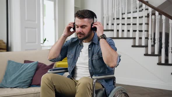 Bearded Guy in Wheelchair which Putting on Earphones and Listening Relaxing Music at Home